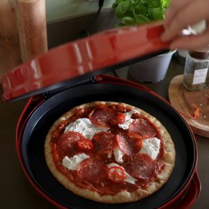 A hand opening a red countertop electric pizza oven to show a fresh pizza cooking inside.