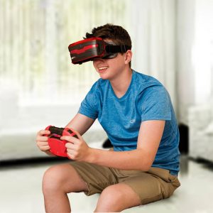 A boy smiling while playing a video game using a red virtual reality headset and steering wheel controller.
