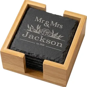 A stack of engraved black slate coasters inside a light wood holder, featuring a custom wedding design that reads "Mr & Mrs Jackson Est. 2026" with floral accents.