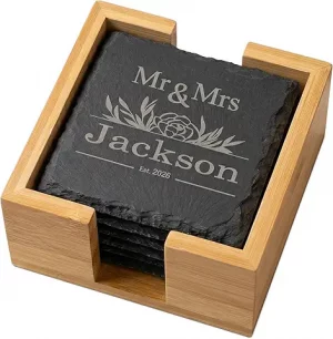 A stack of engraved black slate coasters inside a light wood holder, featuring a custom wedding design that reads "Mr & Mrs Jackson Est. 2026" with floral accents.