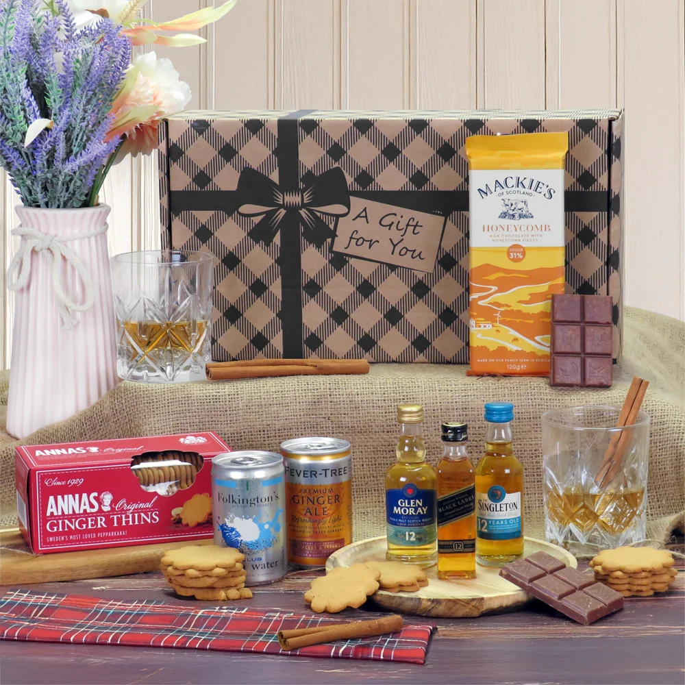 A curated gift hamper box arranged with miniature scotch whisky bottles, chocolate bars, ginger biscuits, mixers, and two glasses.