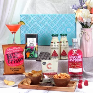 An elegant cocktail and snack gift box set presented on a white marble background. The set highlights a bright pink bottle of Absolut Raspberri, cranberry juice mixers, dark chocolate, chilli crisps, and roasted peanuts set out alongside a prepared raspberry cocktail.