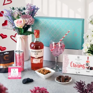 A premium pink gin and chocolate gift box display set against a bright, patterned teal box. The scene highlights Edinburgh Gin Raspberry, pink lemonade mixer, gourmet Whitakers chocolates, Cambrook nuts, and a freshly prepared gin cocktail next to a small bouquet of pink flowers.