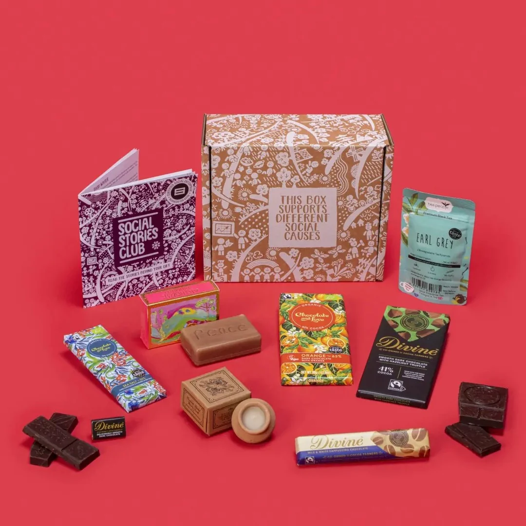 A Social Stories Club ethical gift box against a red background, featuring a patterned cardboard box, a story booklet, Earl Grey tea, Divine chocolates, an organic orange chocolate bar, and a bar of "Peace" soap.