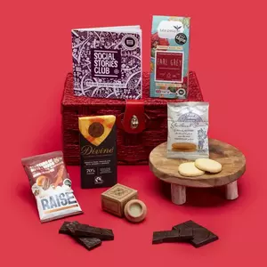 A curated gift hamper from Social Stories Club against a red background, featuring Earl Grey tea, Divine chocolate, shortbread biscuits, Raise maple nuts and seeds, and a small booklet on a red wicker basket.
