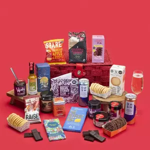 A large Social Stories Club food and drink gift hamper overflowing with gourmet snacks, chocolates, chutneys, spices, coffee, and sparkling water, arranged against a bright red background.