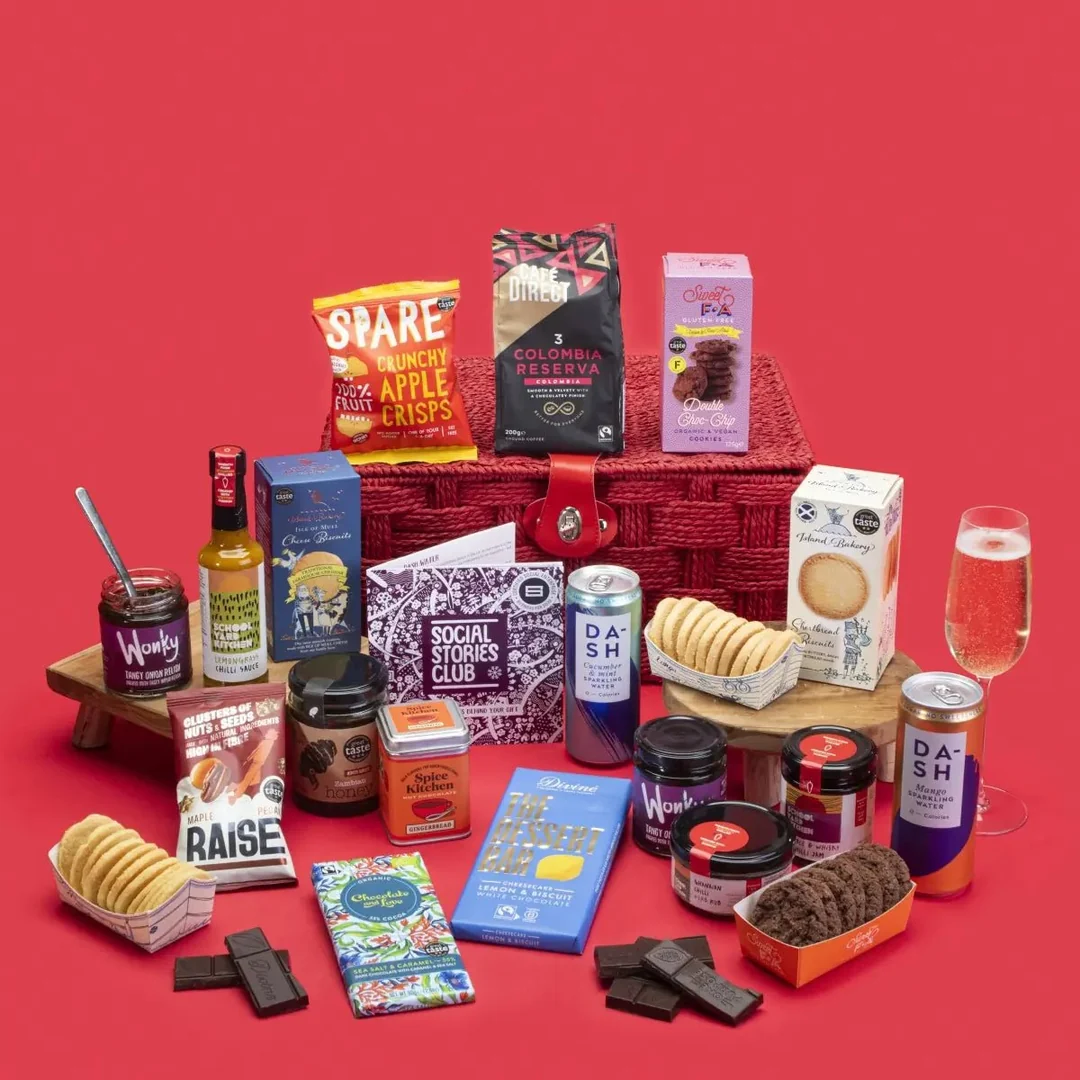 A large Social Stories Club food and drink gift hamper overflowing with gourmet snacks, chocolates, chutneys, spices, coffee, and sparkling water, arranged against a bright red background.