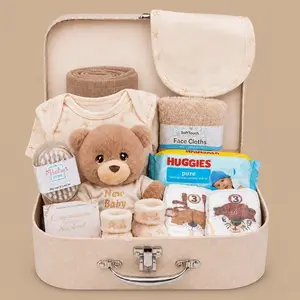 A neutral newborn baby gift hamper arranged inside an open cream vintage suitcase, featuring a plush teddy bear, baby clothes, wipes, and grooming essentials.