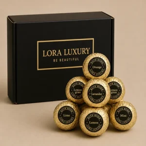 A Lora Luxury Bath Bomb Gift Set featuring a sleek black and gold box next to a small pyramid of gold-wrapped bath bombs with scent labels.