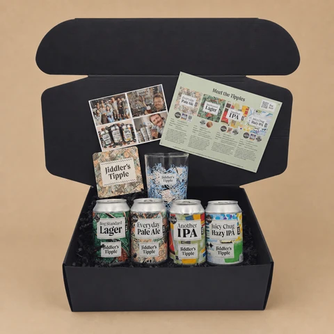 An open black gift box containing a variety pack of four Jiddler's Tipple craft beers with a matching printed glass, beer mat, and tasting guide on a solid beige background