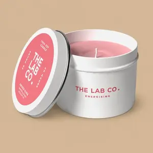 An unlit "The Lab Co. Energising" 100% soy candle with pink wax in a white tin container, next to its matching pink and white metal lid.