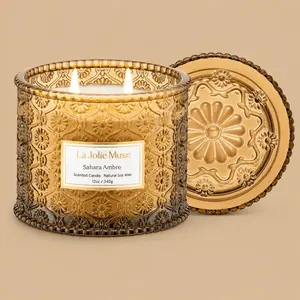 A lit "La Jolie Muse Sahara Ambre" double-wick amber glass jar candle and lid.