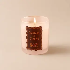 A lit Bloom & Wild "Warm & Calm" candle in a frosted glass holder, radiating a warm flame.