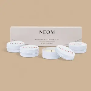 Four NEOM white mini travel candles arranged in front of their sleek, cream-colored scent discovery gift box.