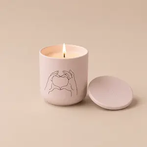 A lit MÆGEN "All The Love" pink ceramic jar candle with a heart-hands illustration MAEGEN Vox All The Love Embers & Musk Candle 240g | ASOS.