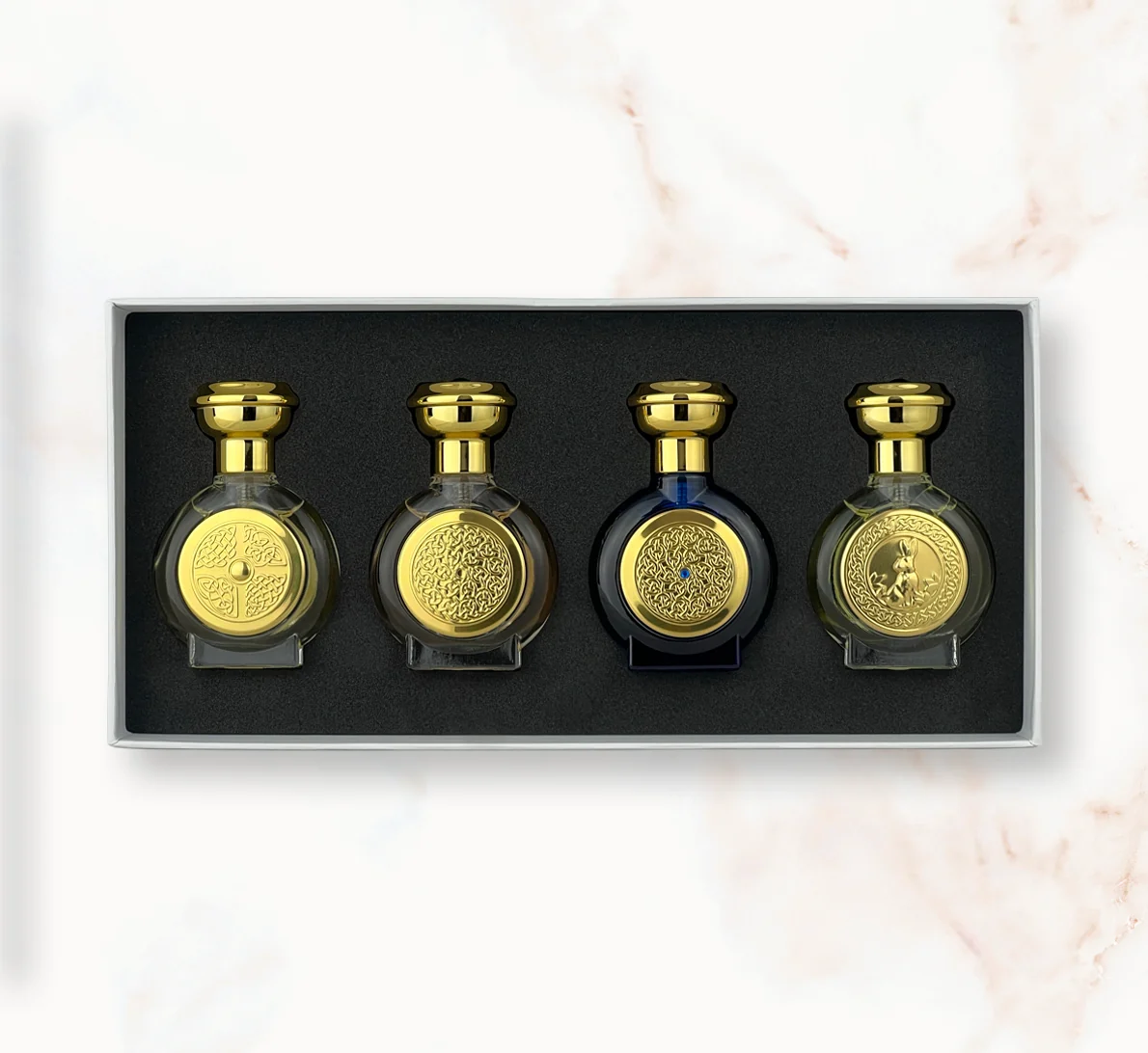 "An open rectangular gift box presenting four miniature, rounded Boadicea the Victorious perfume bottles in translucent olive yellow, clear glass, dark blue, and light translucent green, each styled with a heavy gold cap and a matching circular gold shield emblem."