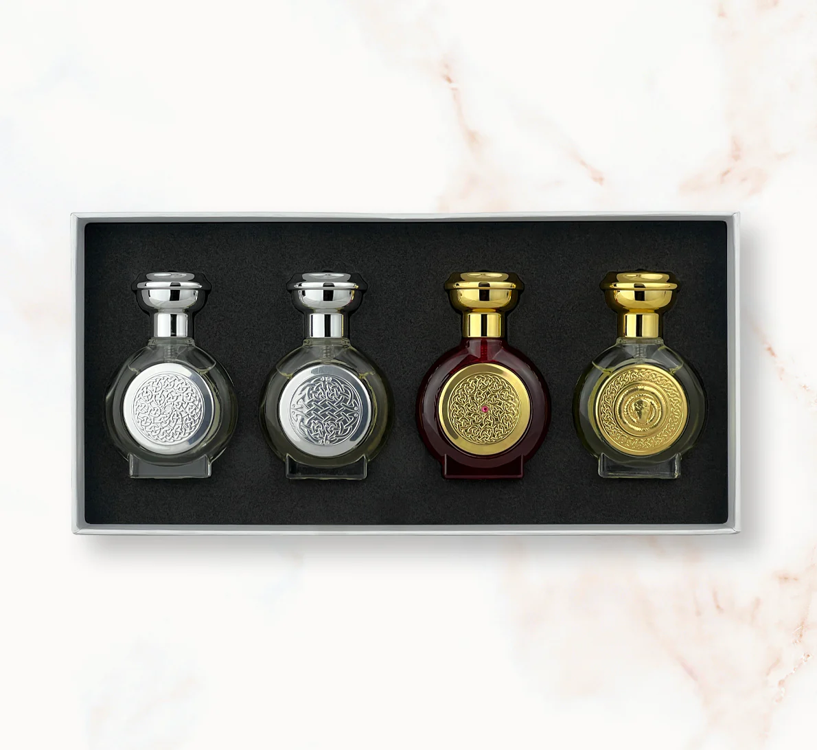 "An open rectangular gift box presenting four miniature, rounded Boadicea the Victorious perfume bottles: two clear glass bottles with silver caps and shields, followed by a dark burgundy bottle and a clear glass bottle with golden caps and shields."