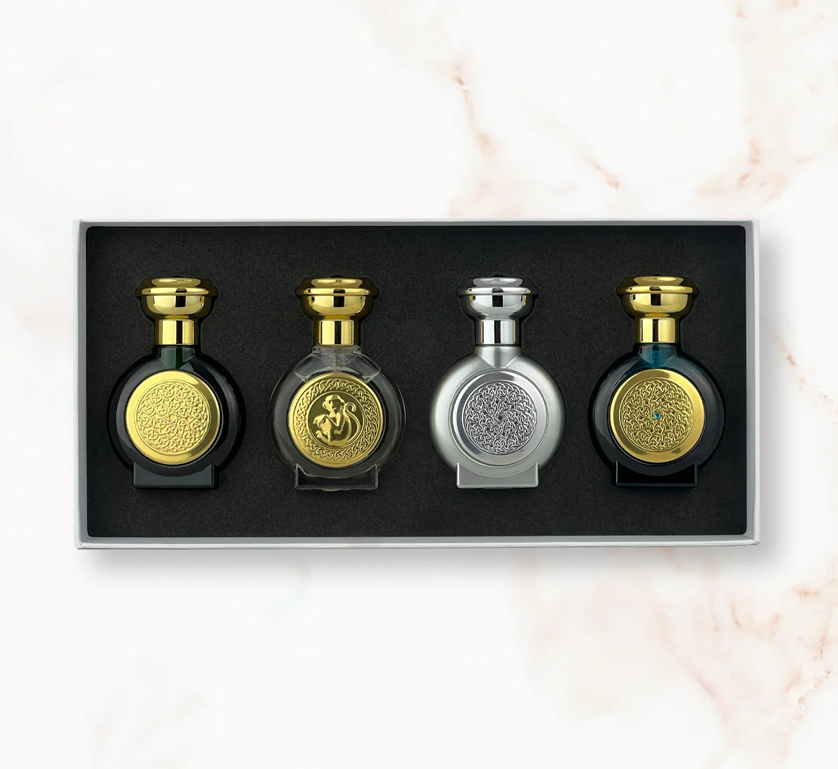 "An open rectangular fragrance gift box featuring four miniature, rounded Boadicea the Victorious perfume bottles in dark green, clear glass with a gold monkey emblem, metallic silver, and deep dark blue, each showcasing detailed circular shields."