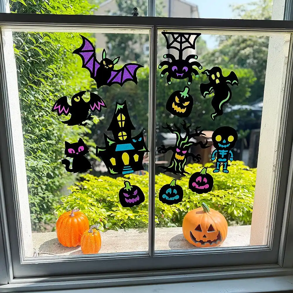 A window decorated for Halloween with colorful gel clings including a haunted house, ghosts, bats, and a skeleton, with three small pumpkins sitting on the window sill.