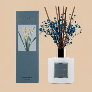 A Cocorrína Cashmere Vanilla reed diffuser set featuring an opaque white glass bottle styled with brown reeds, deep blue baby's breath, and a matching dark teal gift box.
