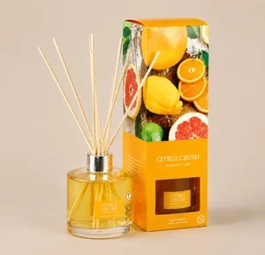 A Wax Lyrical Citrus Crush reed diffuser set featuring a clear glass bottle filled with bright yellow liquid and a vibrant orange gift box decorated with photographic prints of assorted citrus fruits.