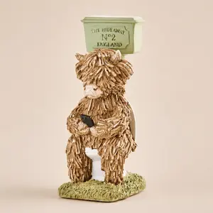 A novelty bathroom ornament of a shaggy Highland cow character using a smartphone while sitting on a high-cistern toilet.