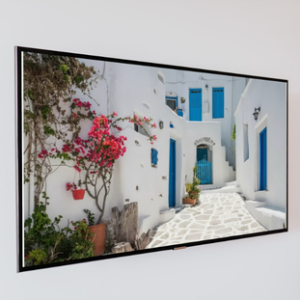A large panoramic framed art print showing a narrow, stone-paved street winding through a traditional Greek village with white-washed buildings, bright blue doors and shutters, and vibrant pink bougainvillea flowers.
