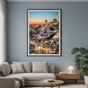 A large vertically framed photography print of a glowing cliffside village in Santorini at sunset, hanging on a blue-gray wall above a modern fabric sectional sofa in a living room.