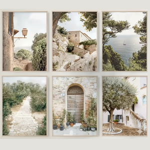 A gallery wall display of six vertical light-wood framed art prints arranged in a two-by-three grid against a light beige wall, featuring warm-toned photography of European coastal landscapes, rustic stone buildings, and olive trees.