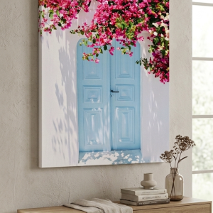 A large vertical canvas art print depicting a light blue double door on a white building surrounded by vibrant pink bougainvillea flowers, hanging above a wooden console table.