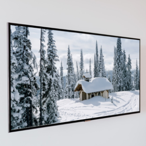 A large horizontal framed photograph of a snow-covered small cabin surrounded by pine trees in a winter forest, mounted on a plain white wall.