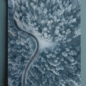 A vertical wall art print hanging from a white wooden hanger bar on a dark blue wall, showing an aerial top-down view of a winding road cutting through a snow-covered pine forest.