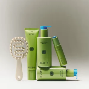 A lineup of five Base-K hair care products in sage green packaging arranged with a beige scalp massage brush against a neutral gray background.