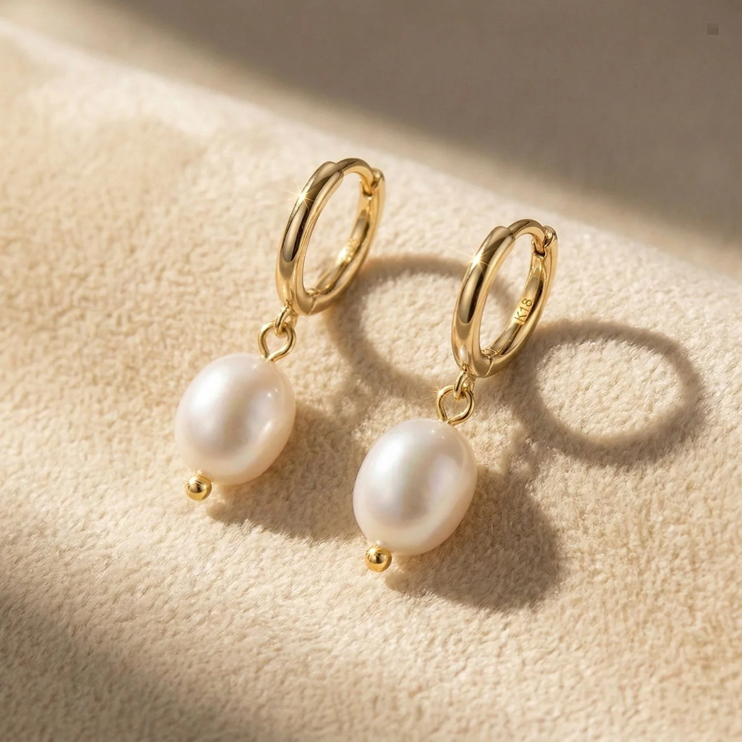 Gold huggie hoop earrings with hanging white pearl drops resting on a cream velvet surface.