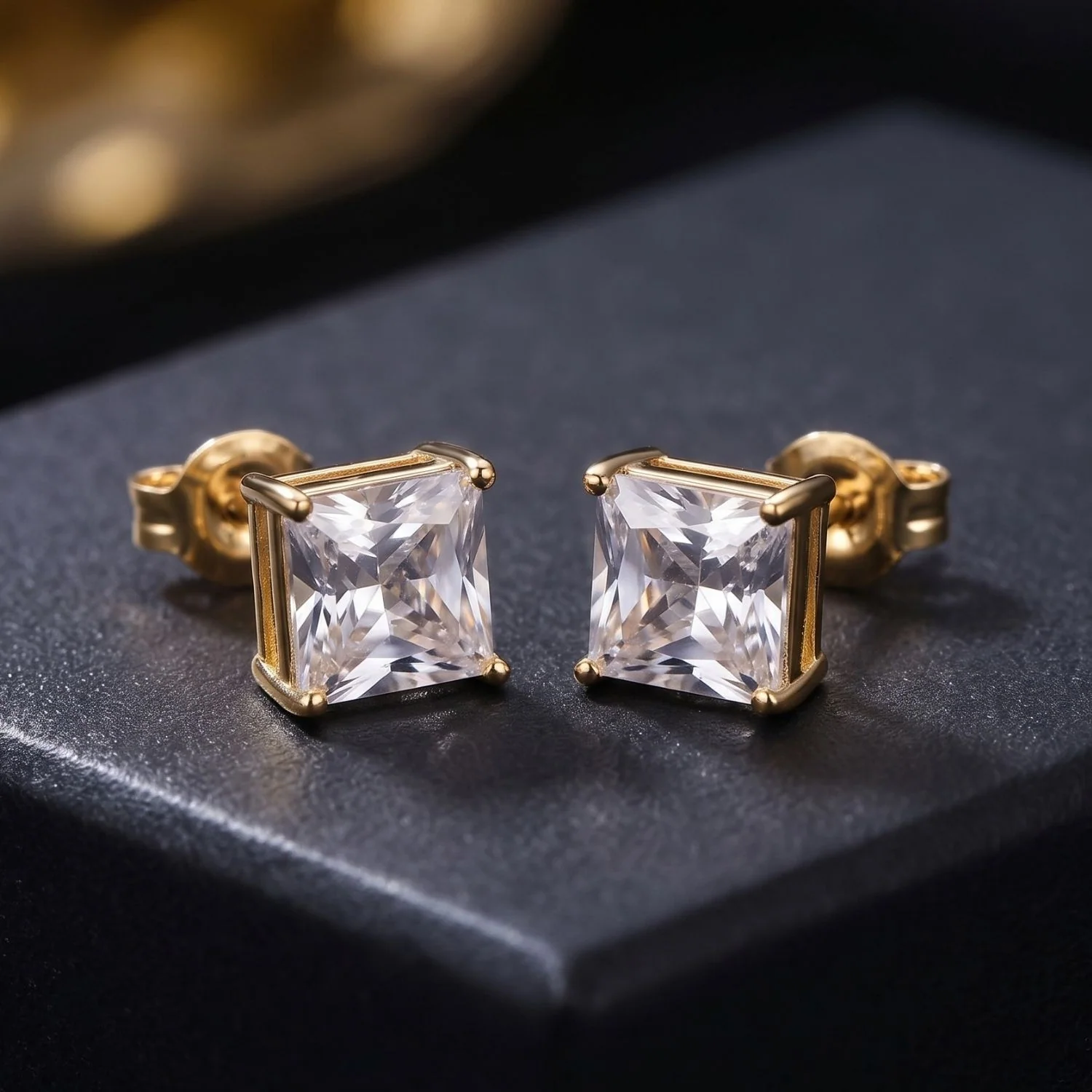 A pair of classic yellow gold solitaire stud earrings featuring sparkling princess-cut square gemstones, displayed on a dark grey jewelry box.