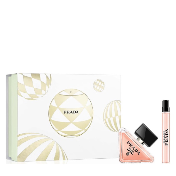 Prada Paradoxe fragrance gift set with a white box featuring gold holiday bauble ornaments, a triangular pink perfume bottle, and a matching slim travel spray vial.